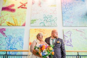 A couple poses together against a vibrant wall with colorful artwork featuring bright yellow, blue and pink patterns.