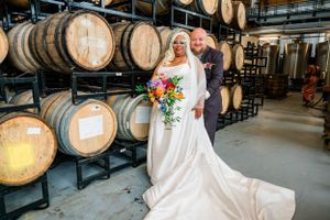 A wedding couple poses in front of stacked oak wine barrels inside a rustic winery cellar.