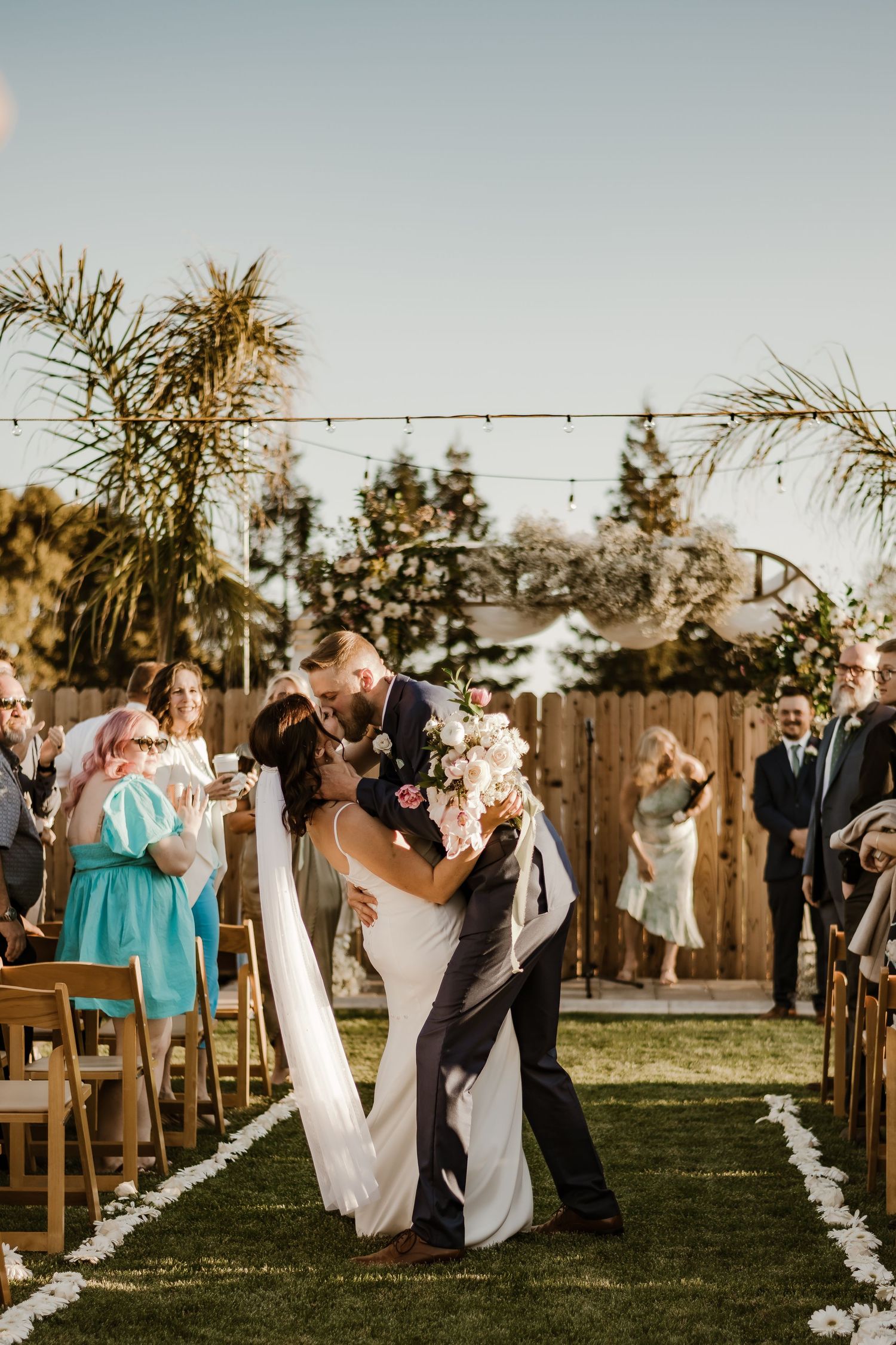 Outdoor wedding ceremony with guests seated on wooden chairs along a petal-lined aisle.