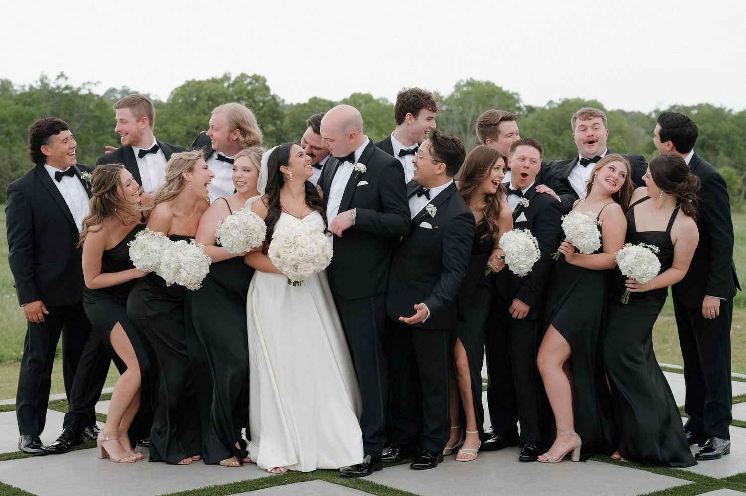 Wedding party in formal black attire and white bridal gown gather together for fun group photo outdoors.