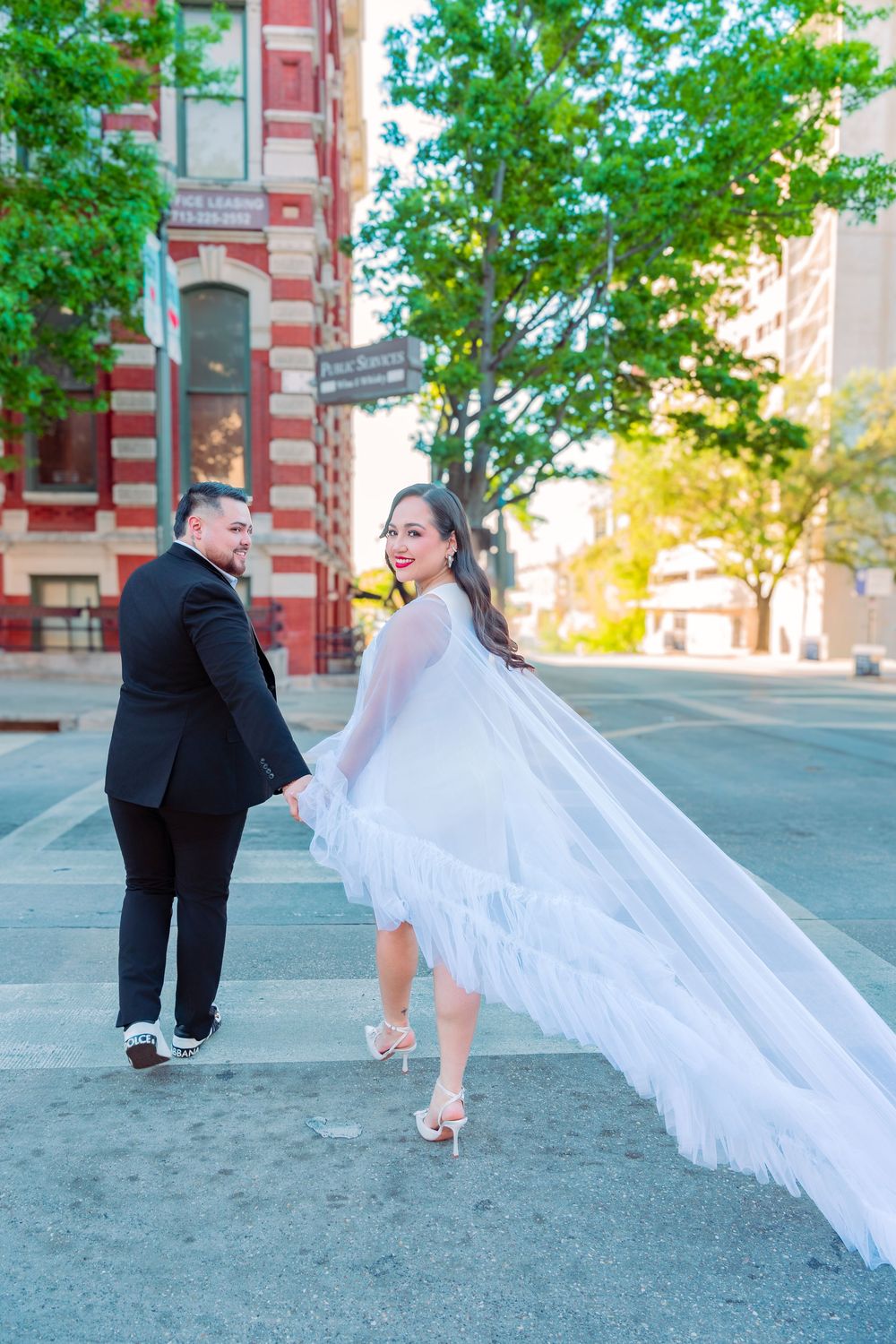 Bride in flowing white cape dress holds groom's hand while looking back smiling on a sunny urban street.