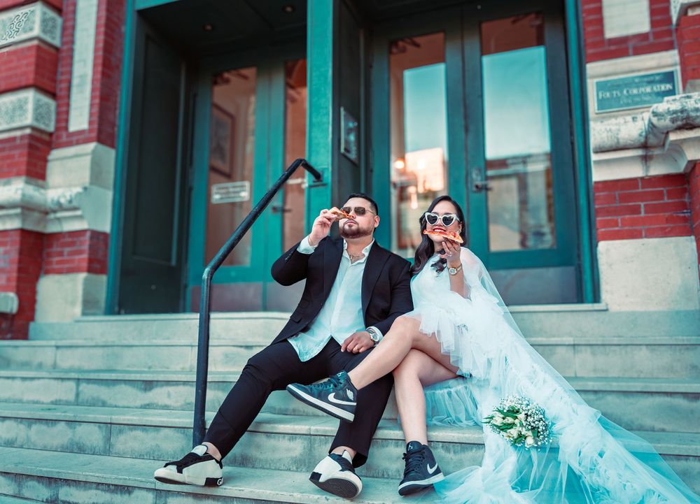 Bride and groom sitting on steps in wedding attire with sneakers, blowing bubbles outside a brick building.