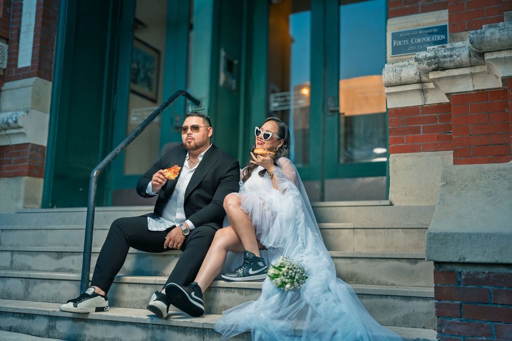 Bride and groom sitting on building steps eating, wearing sunglasses, with wedding dress and black suit with sneakers.