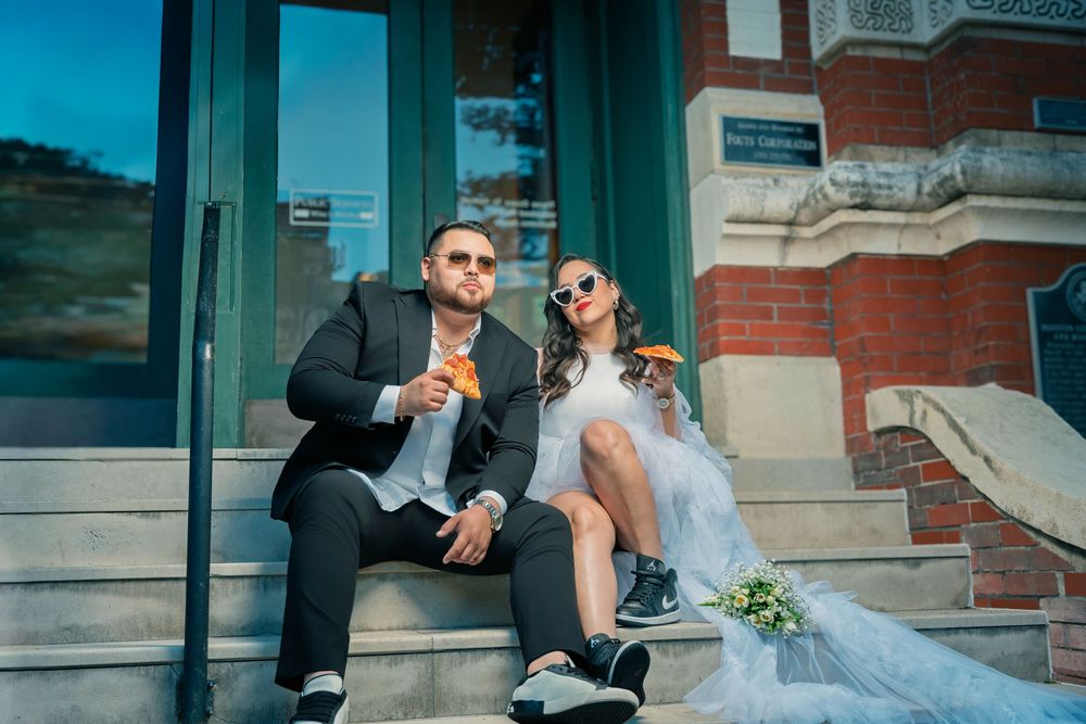 Stylish bride and groom sitting on building steps in sunglasses, enjoying snacks in wedding attire with sneakers.