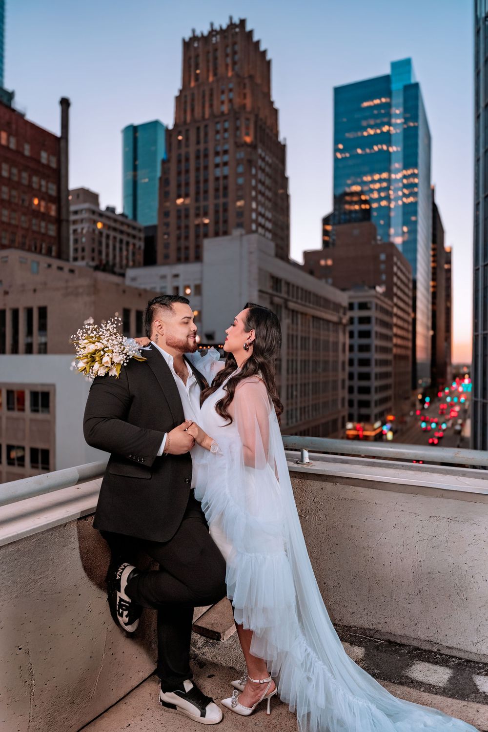Newlywed couple poses romantically on rooftop with stunning city skyline at sunset behind them.