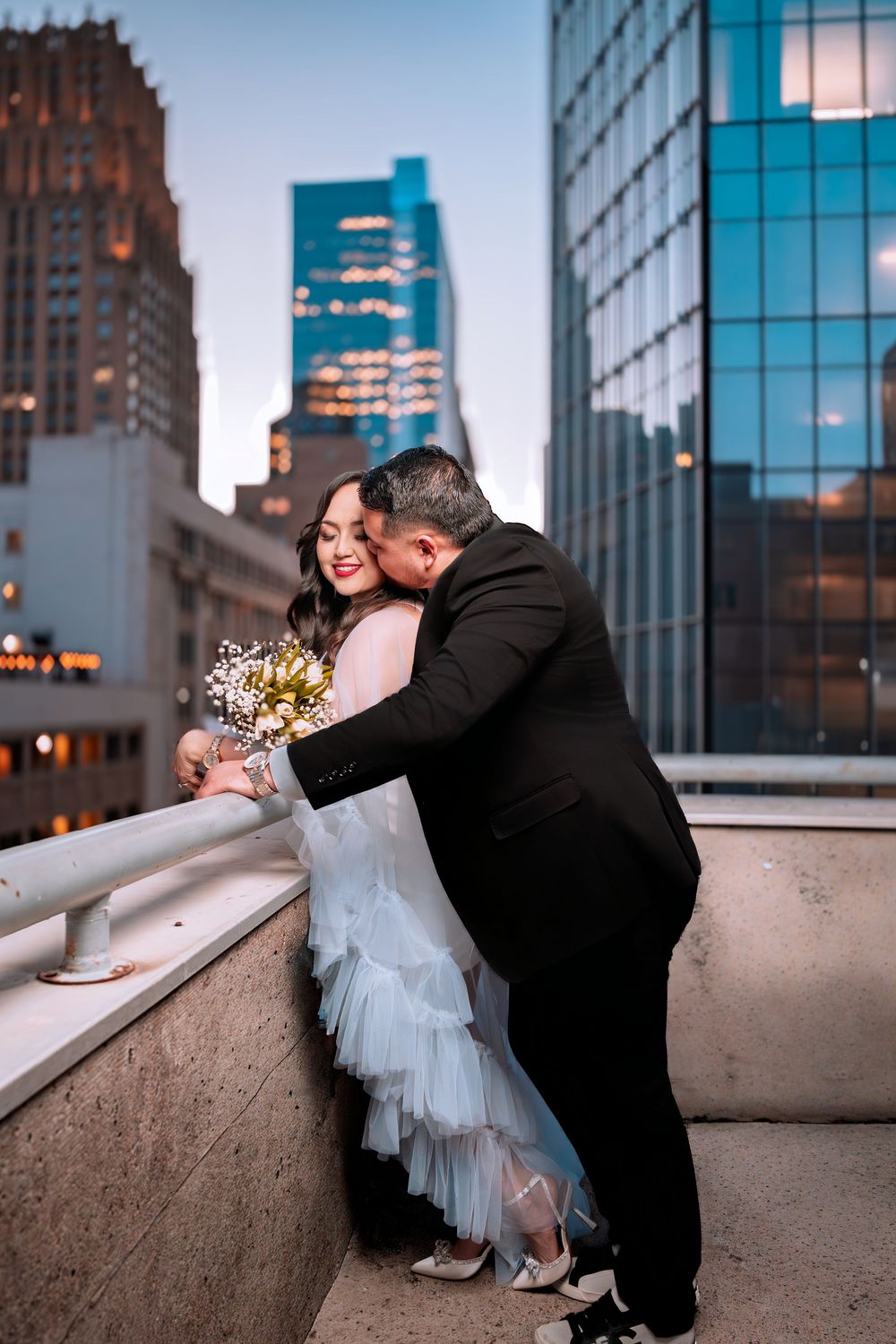 Groom in black suit embracing bride in layered tulle dress on urban rooftop at dusk with city skyline.