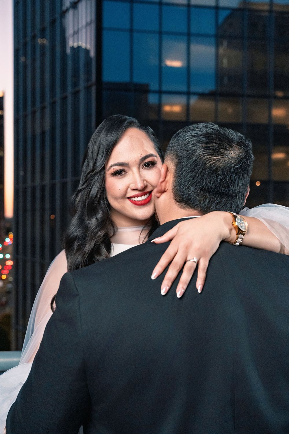 Smiling bride in white dress embraces groom on rooftop at dusk, showing engagement ring with city skyline behind.