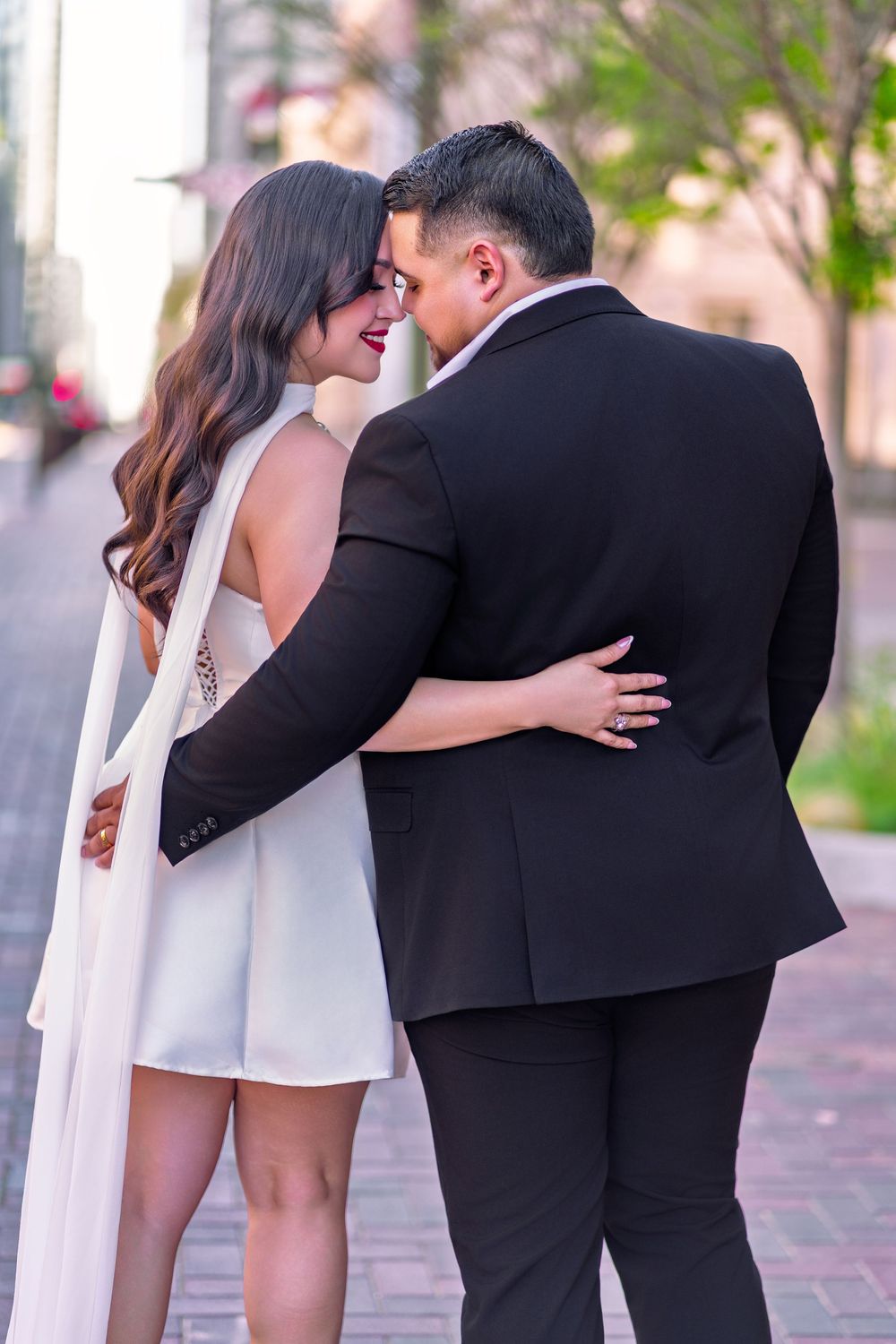 Couple in formal wear embracing and smiling on a brick walkway, woman in white dress, man in black suit.