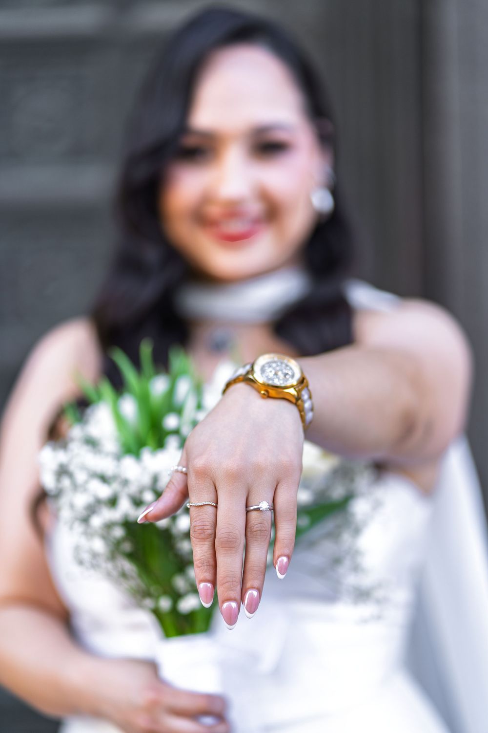 Smiling bride in white dress shows off wedding ring and gold watch while holding white floral bouquet.