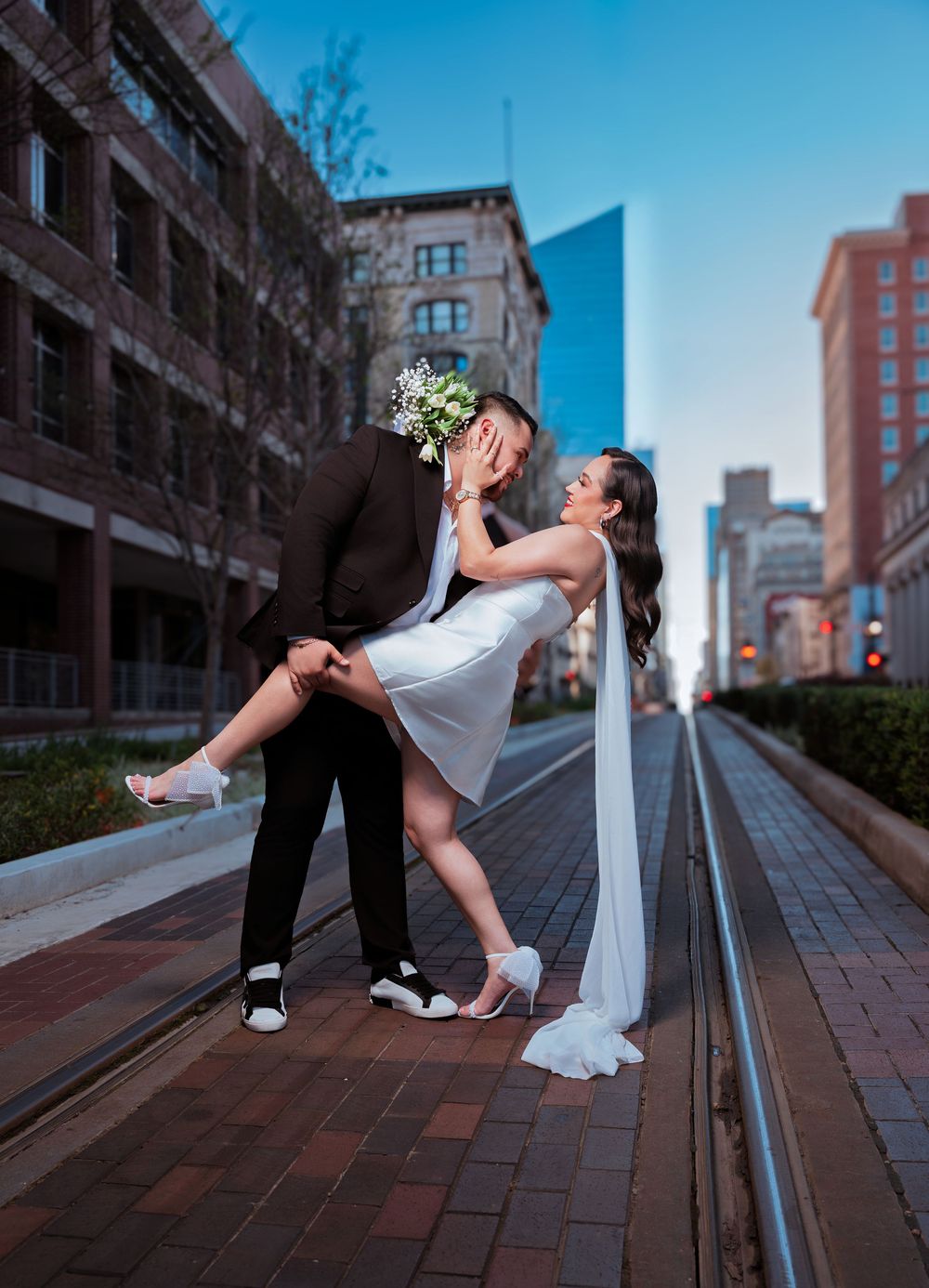 Bride and groom share a romantic dip kiss on urban street railway with city buildings backdrop at dusk.