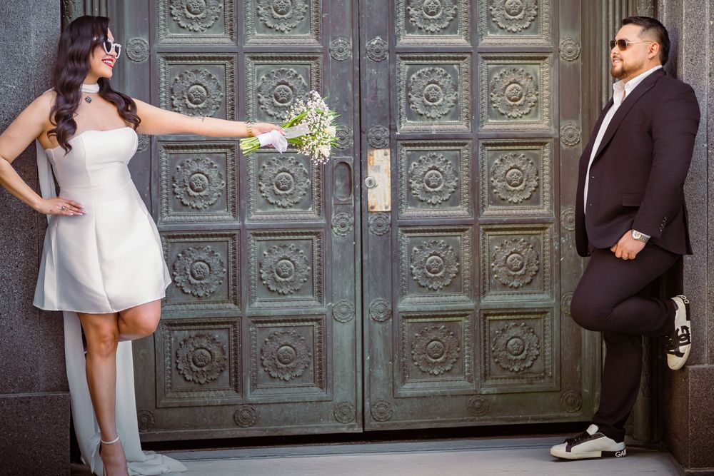 Bride in white mini dress holds bouquet toward groom in black suit, posing before ornate vintage doors.