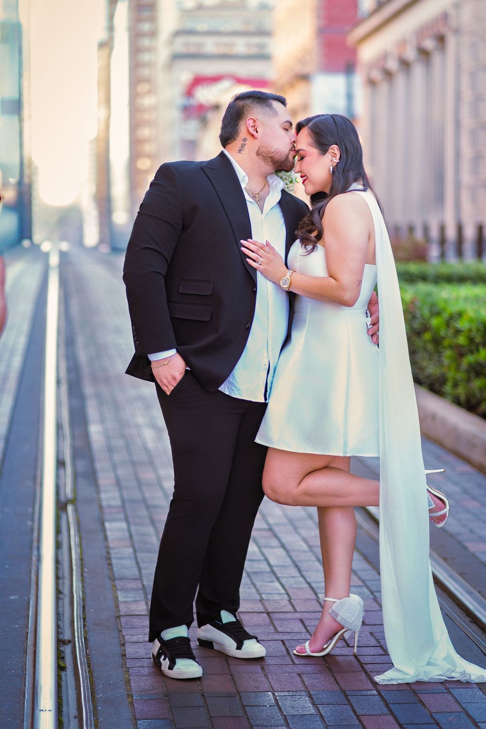 Couple sharing a romantic kiss outdoors, woman in white mini dress with cape, man in black suit, urban street backdrop.
