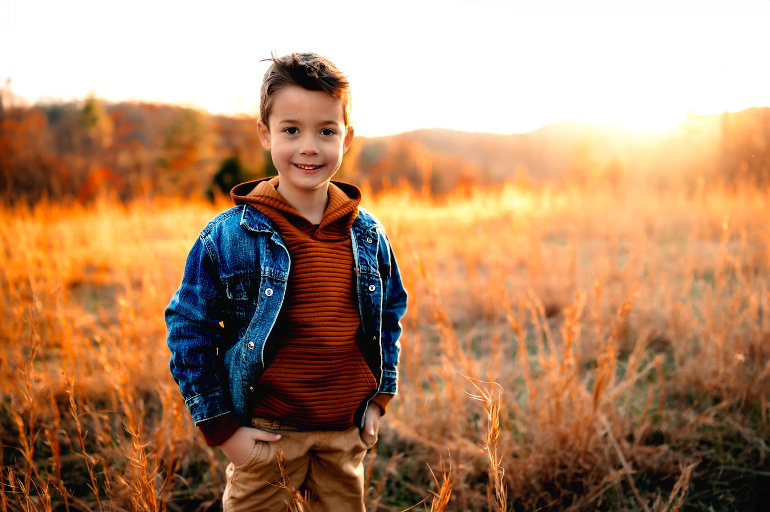 Young child in denim jacket smiling in golden autumn field during sunset.