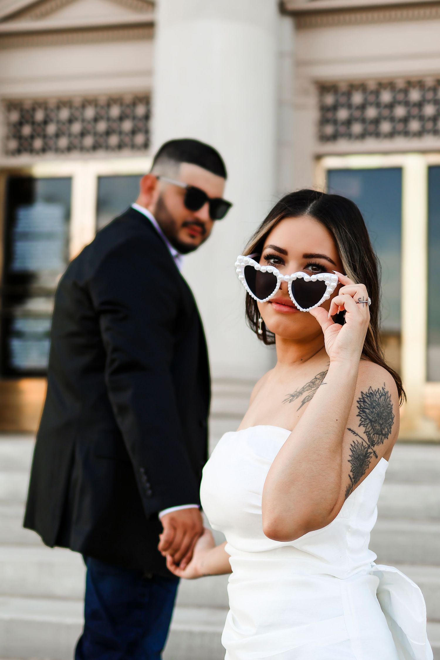 Stylish couple in formal wear posing with heart-shaped sunglasses near building columns.