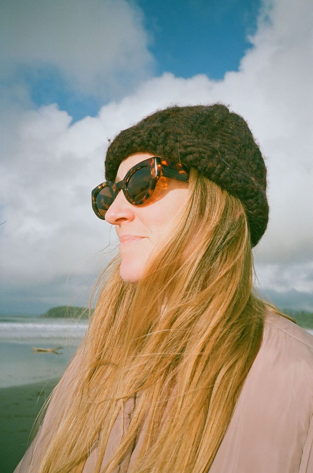 Blonde woman in black knit beanie and sunglasses smiling on a cloudy beach.