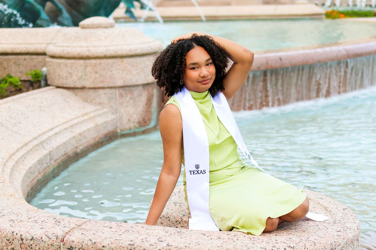 Person in light green dress poses gracefully by curved fountain on sunny day.