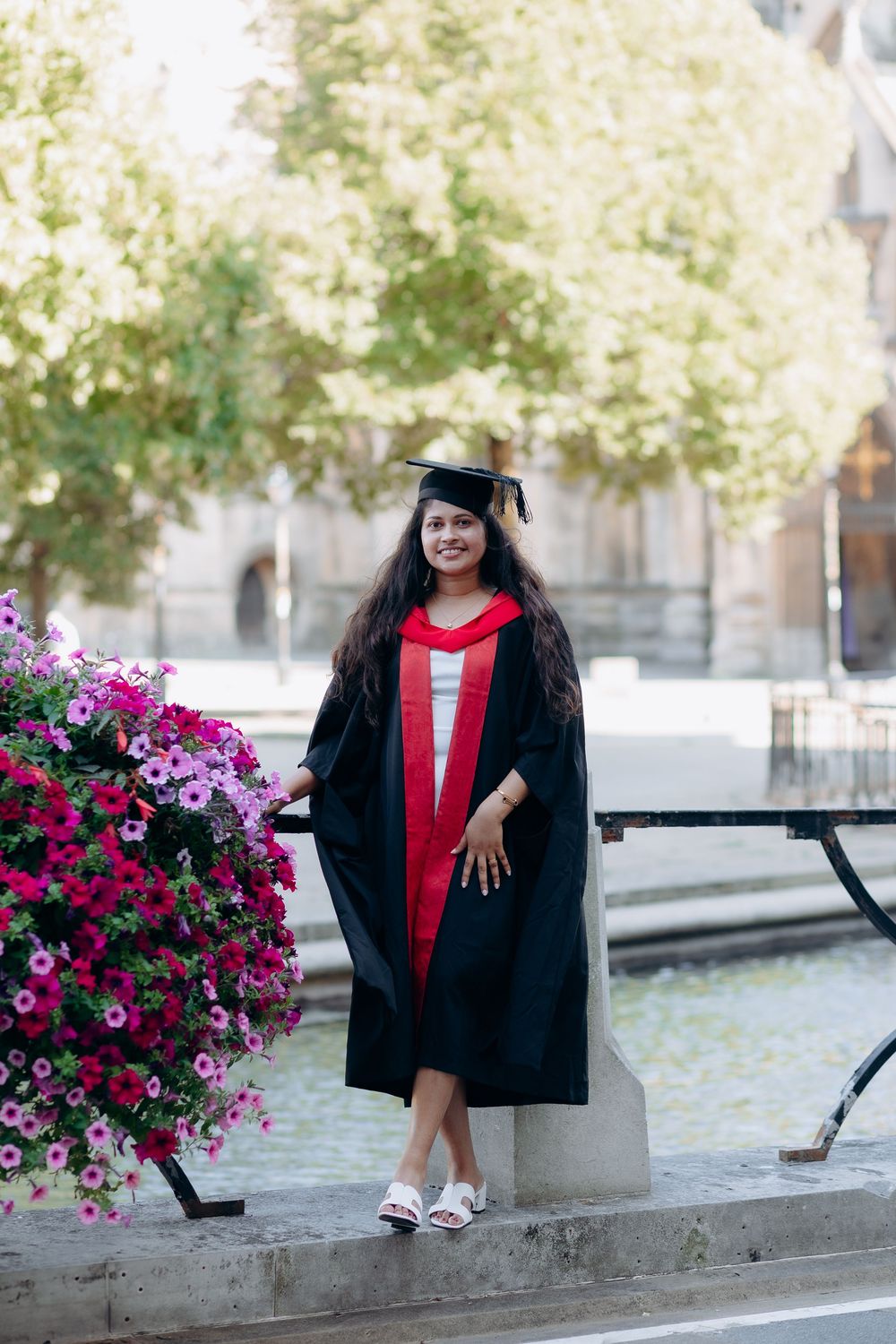 Graduate in academic regalia poses near vibrant pink flowers during outdoor graduation photo session.