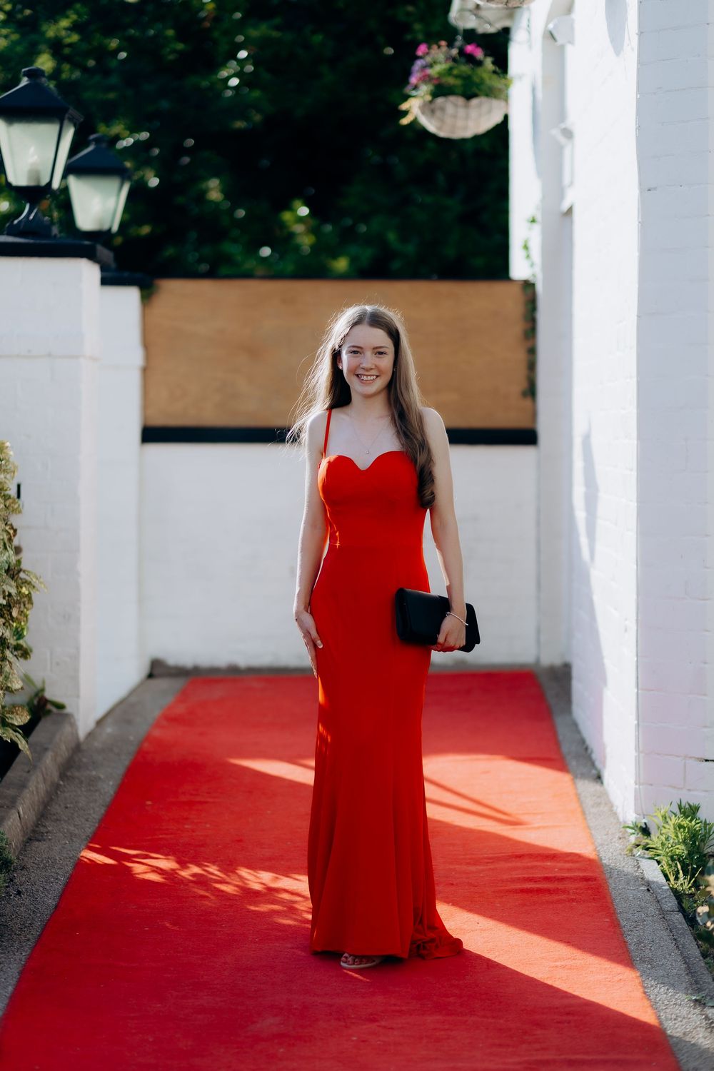 A person in a red formal dress poses on a red carpet walkway framed by white pillars in an outdoor setting.