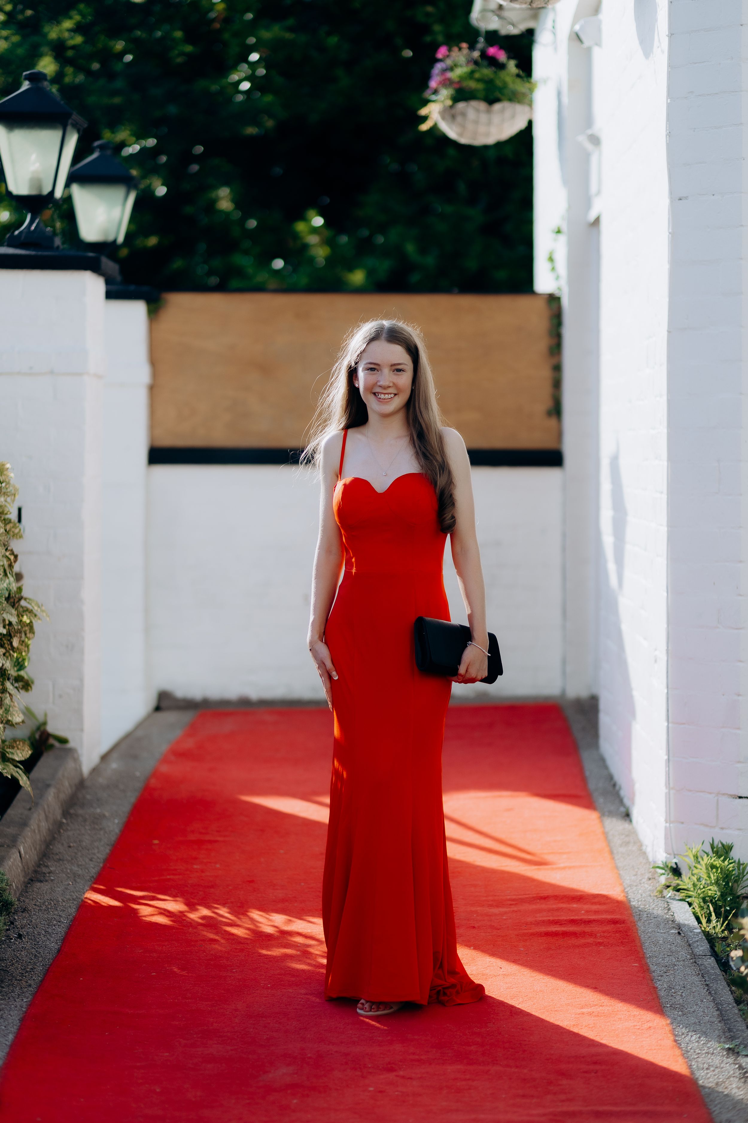 A person in a red formal dress poses on a red carpet walkway framed by white pillars in an outdoor setting.