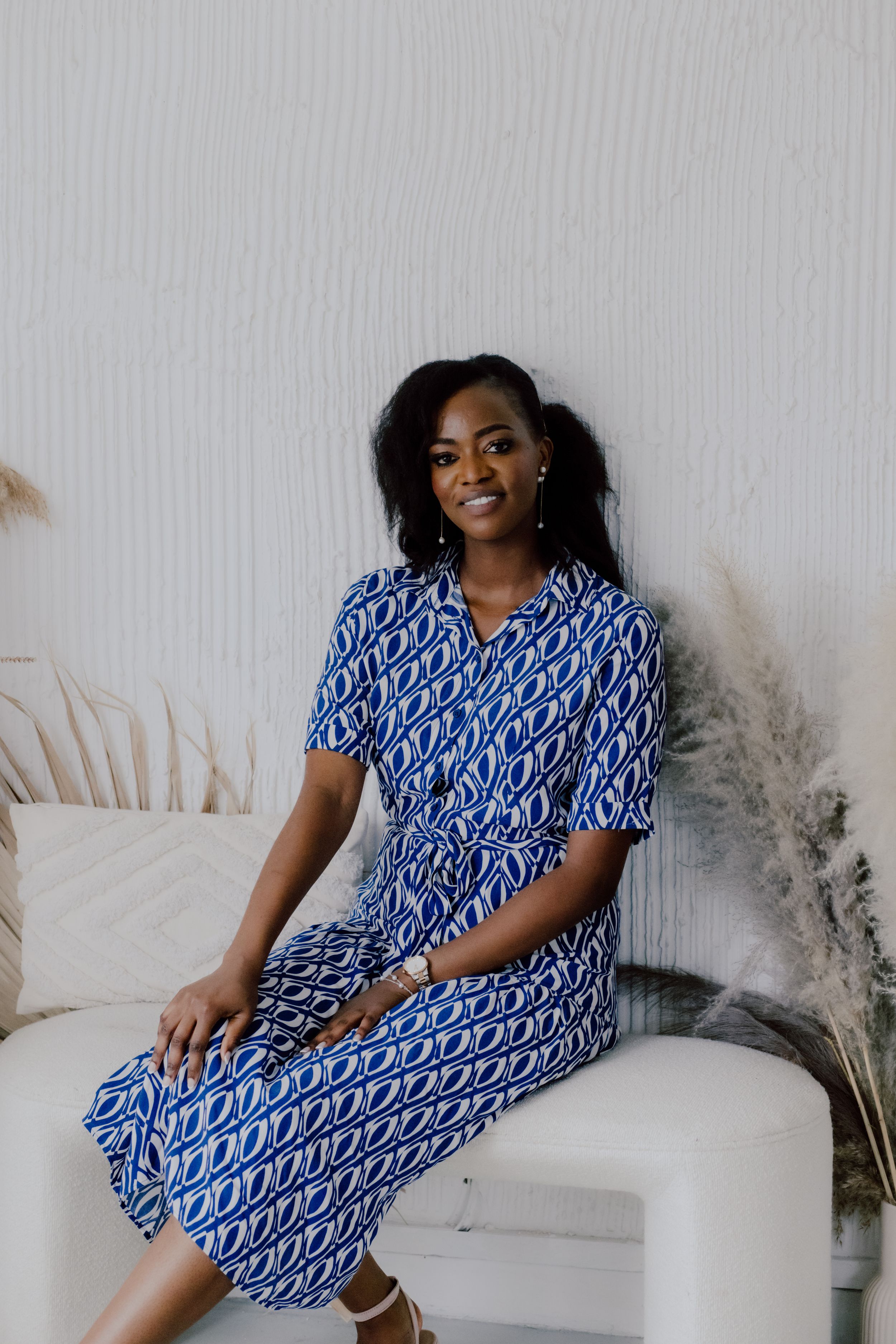 Woman in stylish blue patterned dress poses elegantly on white sofa with decorative pampas grass.