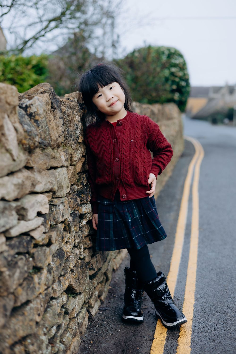 A small child in a burgundy knit sweater and denim stands near a stone wall on a road with yellow lines.