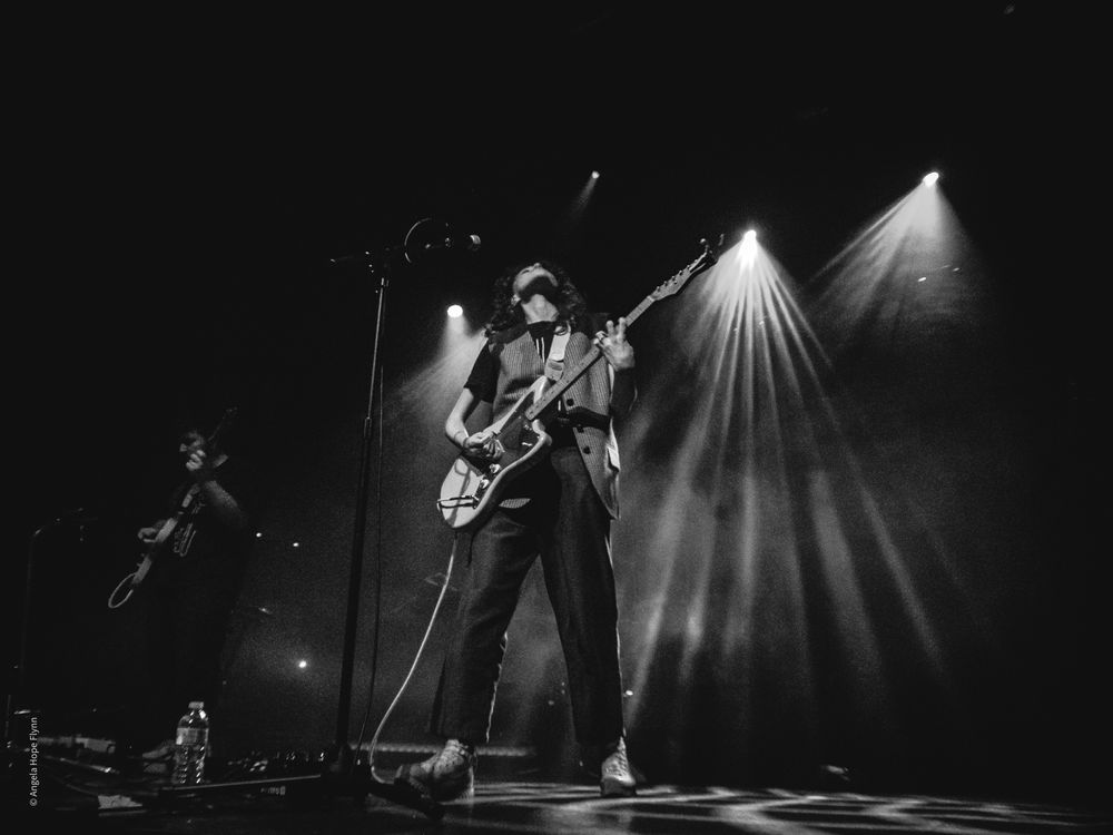A performer plays guitar on stage in dramatic black and white lighting during a concert.