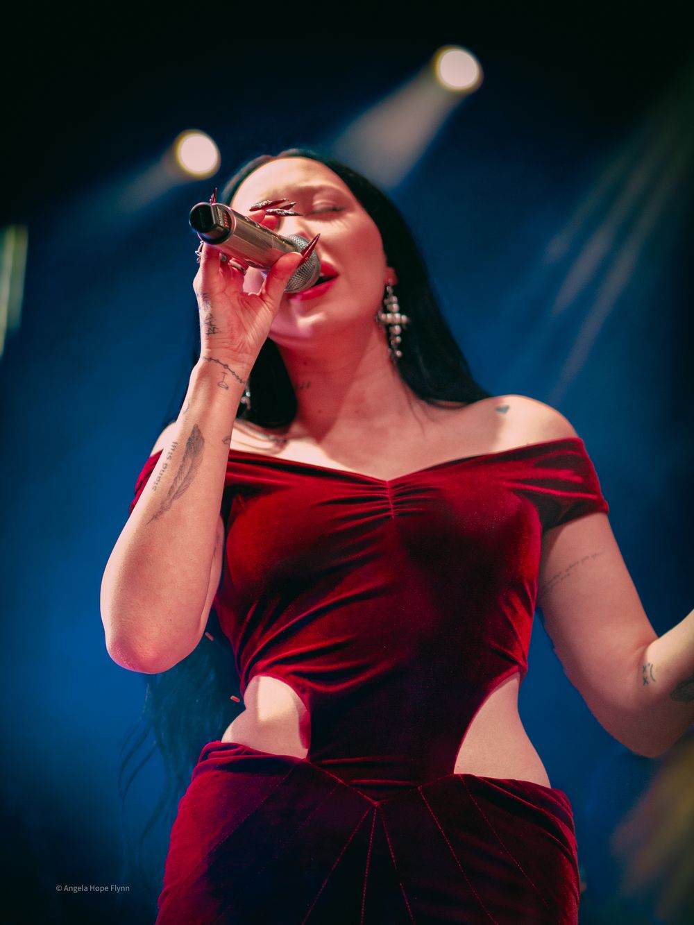 A performer in a red velvet off-shoulder outfit sings passionately on stage under dramatic blue lighting.