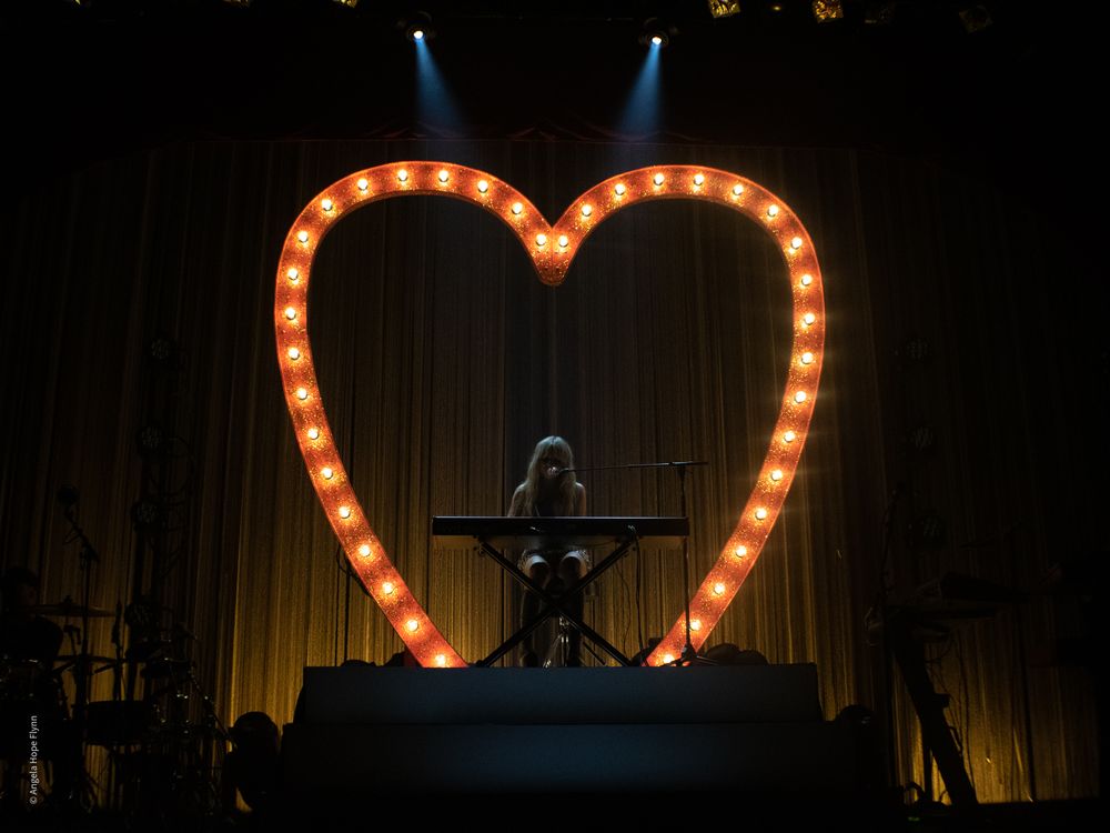 Performer silhouetted against large illuminated heart prop on dark stage.