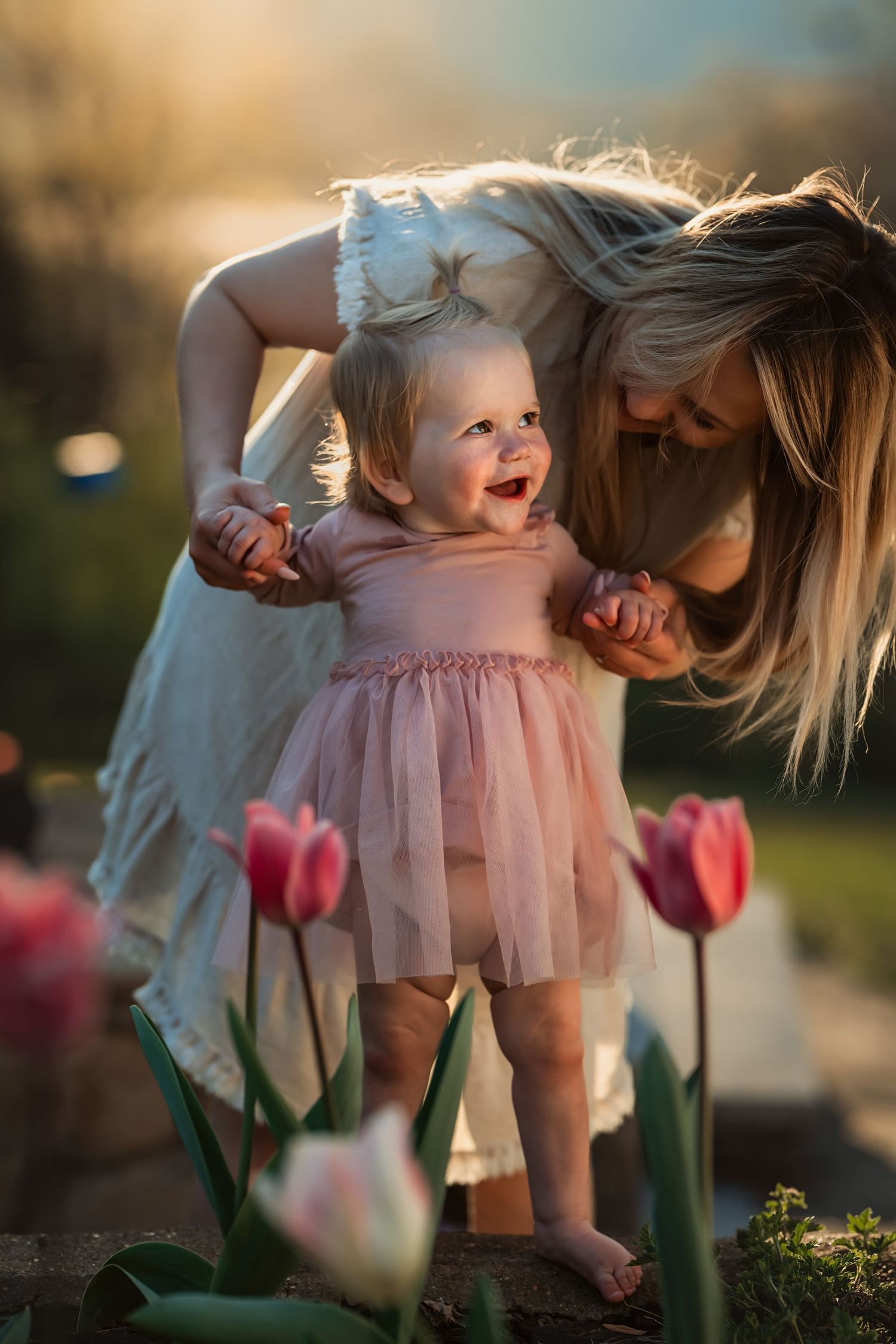 mom holds baby daughter in pink so she can stand behind some tulips
