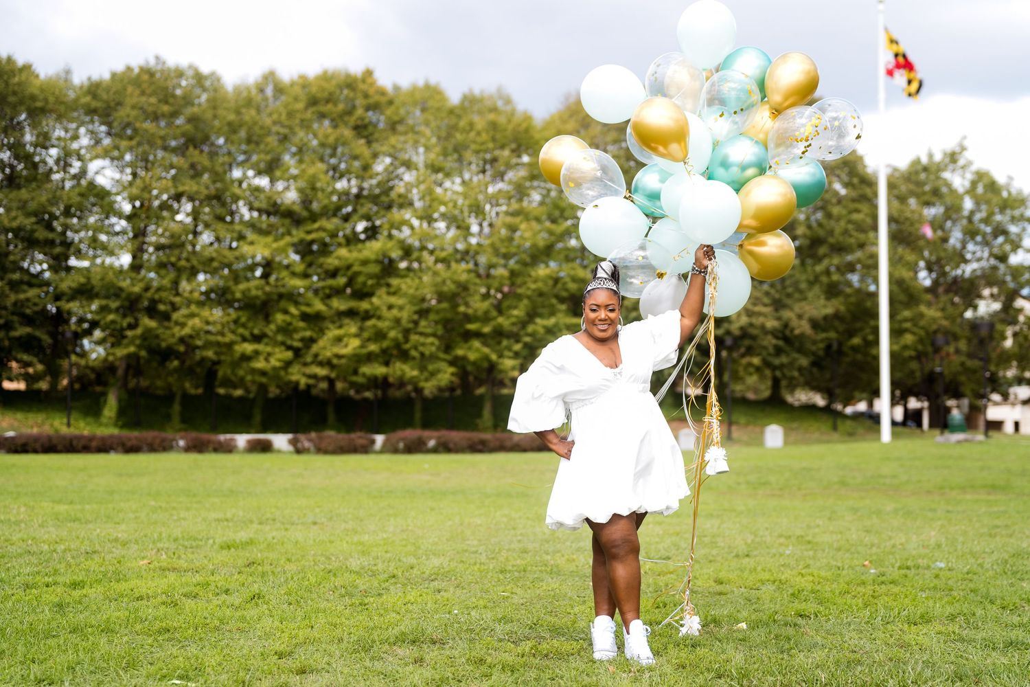 A person in white holds colorful balloons while standing on green grass with trees in the background.