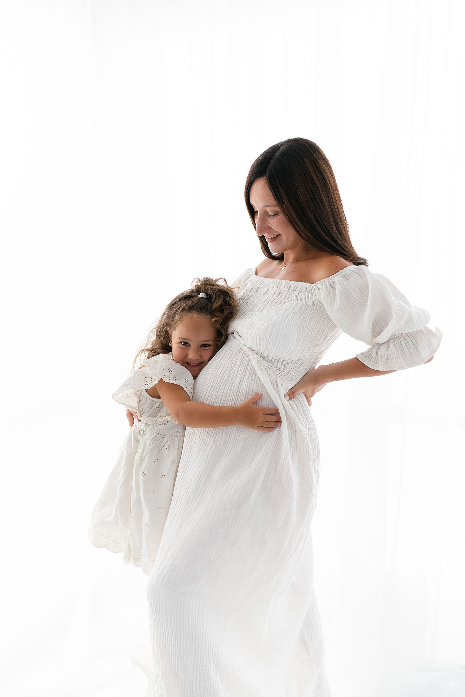 Mother and daughter in matching white dresses share tender moment during maternity photoshoot.