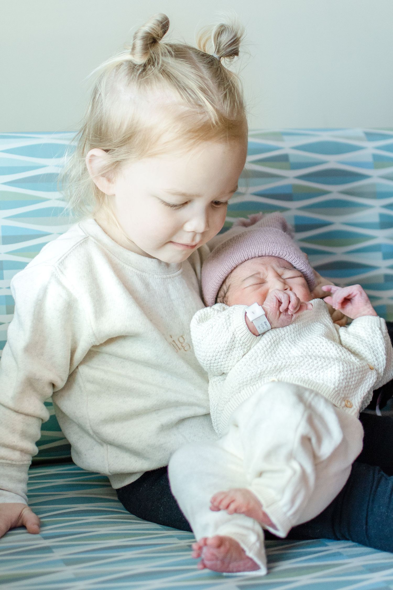 Toddler in white sweater holding and looking down at newborn baby on blue patterned couch.