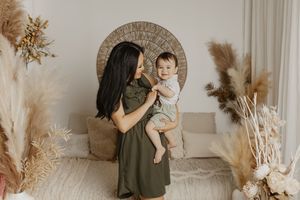 Mother in olive dress holds smiling baby in bright boho studio with pampas grass and warm neutral decor.