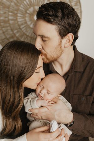 A tender family moment as parents share gentle kisses with their newborn baby in a warm-toned studio portrait series.