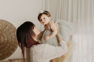 A mother in a lace dress lifts her baby girl in a green tutu dress, sharing a tender moment indoors.