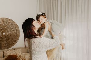 A mother in a white lace dress lifts and kisses her baby girl in a tulle dress in a bright, bohemian room.