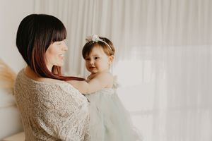 A mother in a lace top holds her toddler daughter in a white dress and floral headband near a bright window.