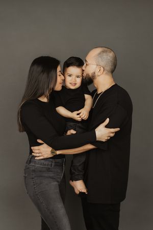 Family of three in matching black outfits, parents kissing toddler on cheeks in studio portrait.