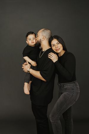 Family of three in matching black outfits posing for a studio portrait, father kissing baby while mother smiles.