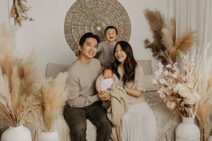 Happy Asian family of four poses in boho studio with pampas grass, parents holding newborn baby and toddler smiling.