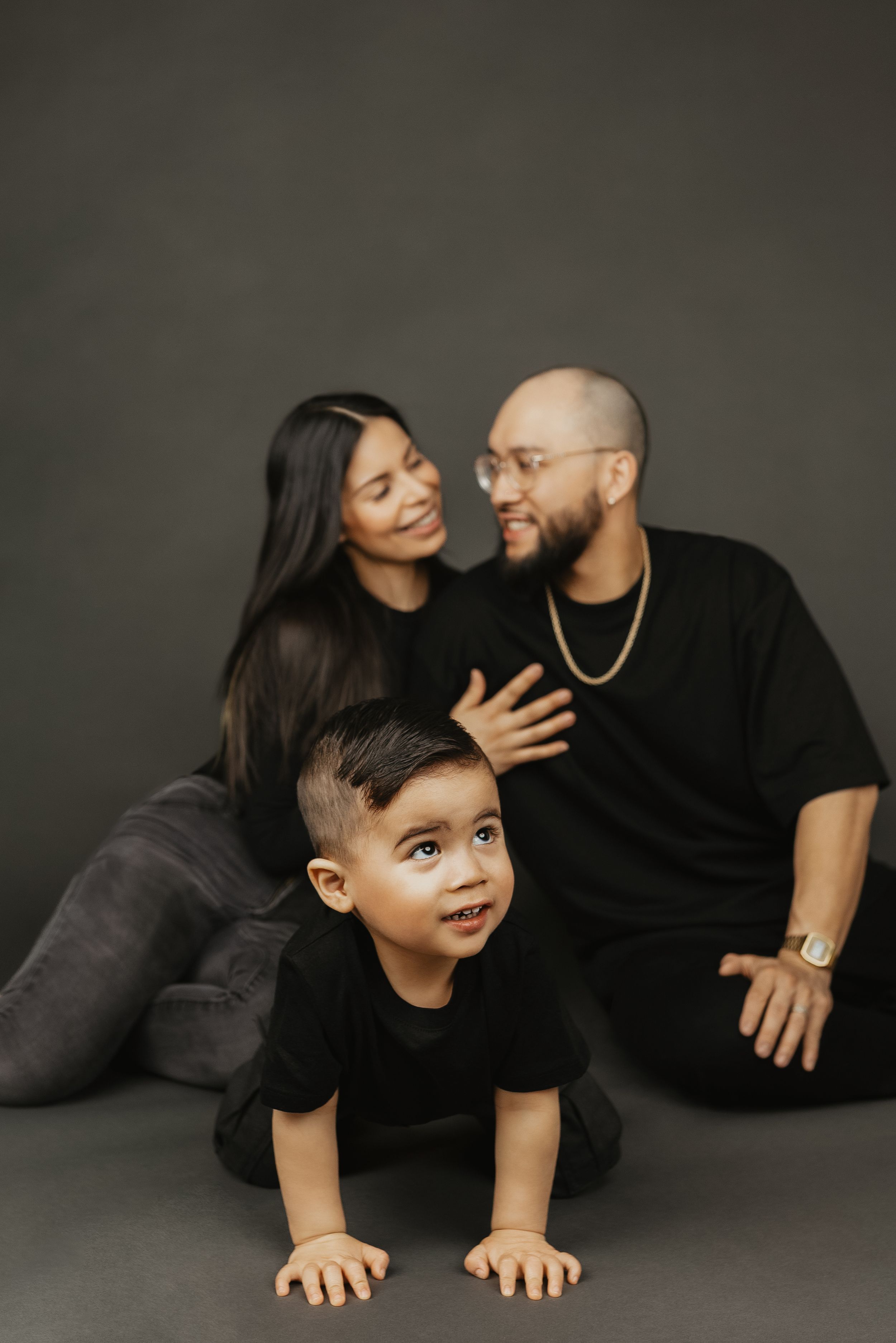 Family photo with parents in black sitting on floor while toddler crawls forward in studio.
