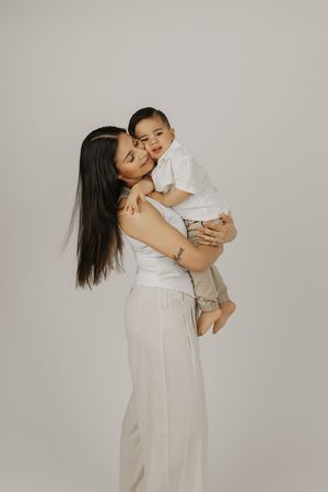 Mother in white outfit holding and kissing her baby in a bright studio portrait session.