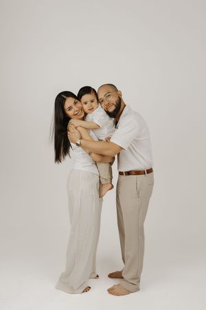 A happy family in neutral tones, parents embracing their young child while posing barefoot in a bright studio.