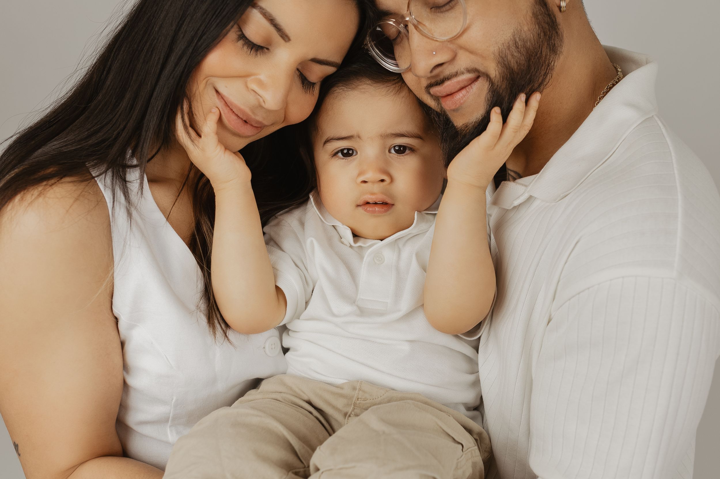 A young family poses together, parents in white shirts tenderly holding their baby boy in a soft studio portrait.