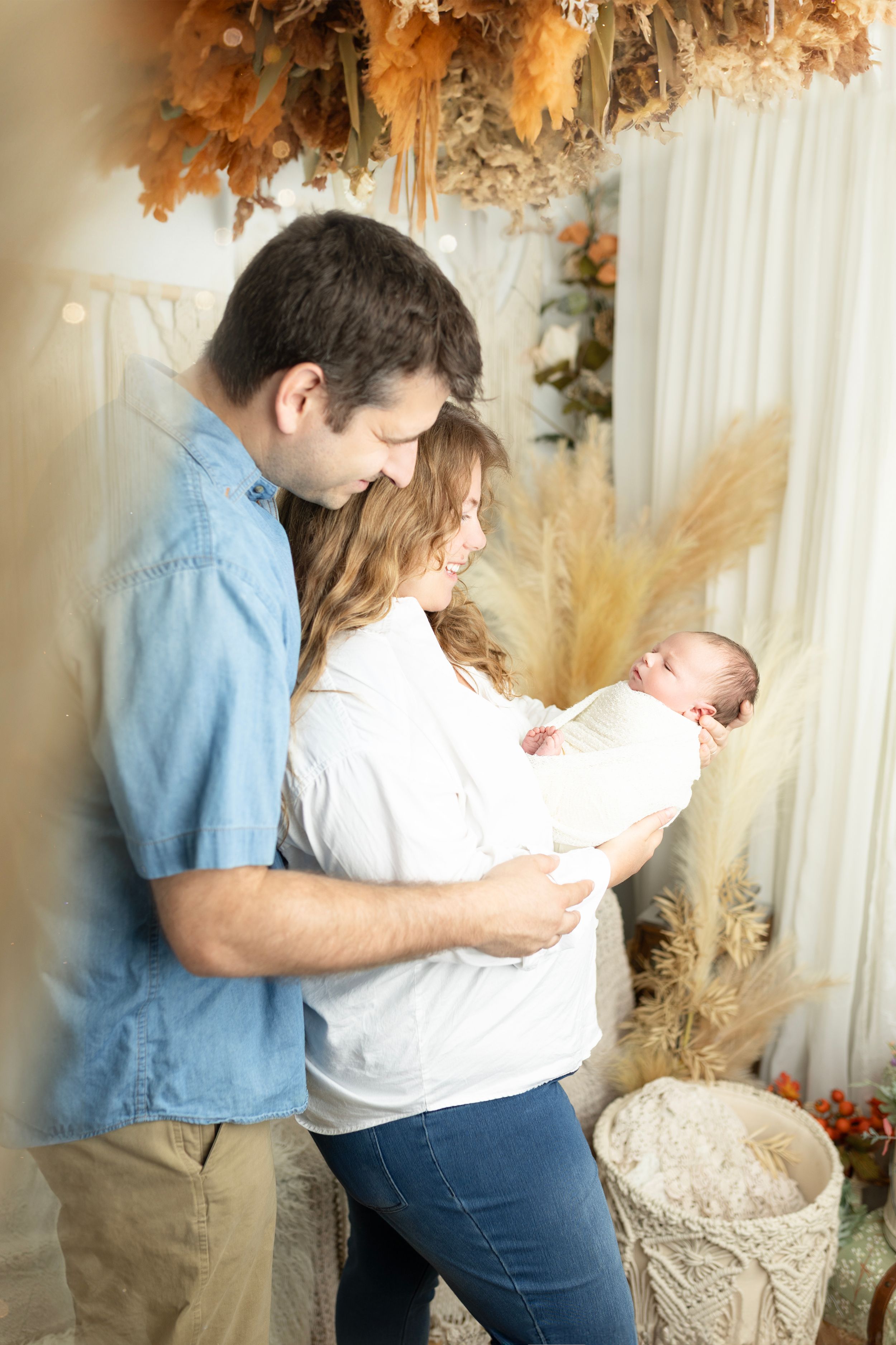 A couple shares tender moments with their newborn baby against an autumn-themed backdrop with warm lighting.