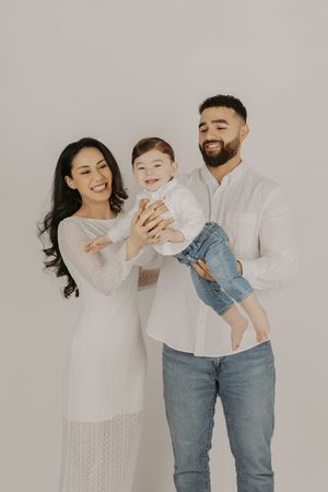 Happy family portrait with mother, father, and smiling baby dressed in white and denim against a light background.