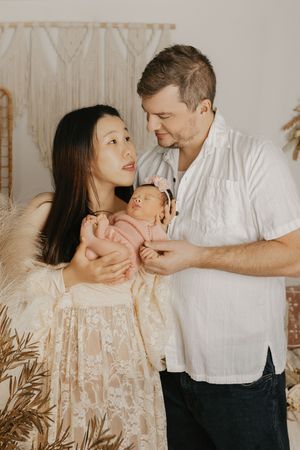 A couple lovingly holds their newborn baby in front of a beige backdrop with dried pampas grass decor.