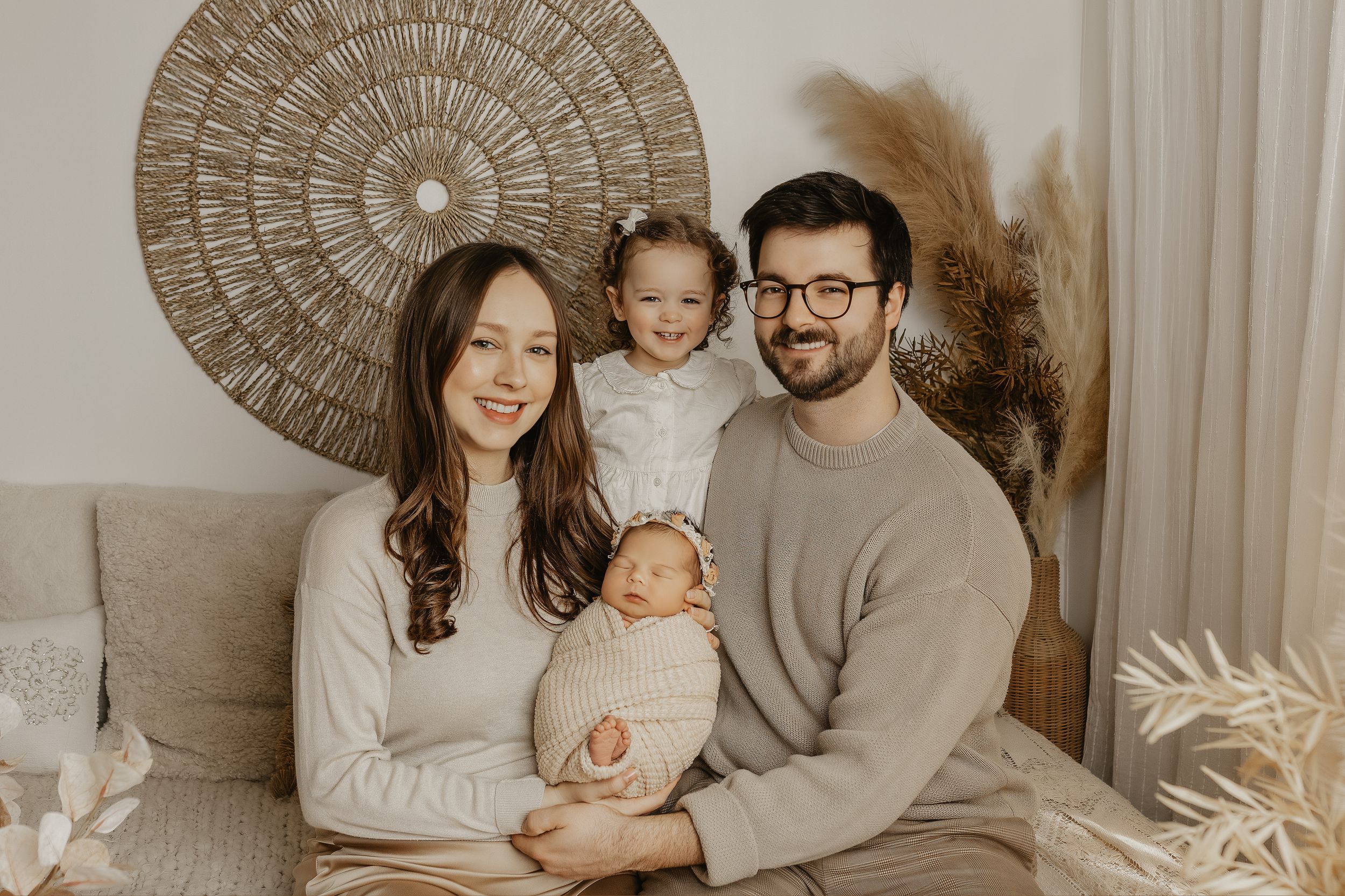 Happy family of four posing for a professional newborn photo session in a cozy, neutral-toned boho studio setting.