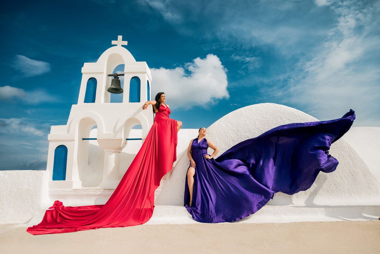 Two flowing dresses in red and purple create drama against a white church in Santorini.