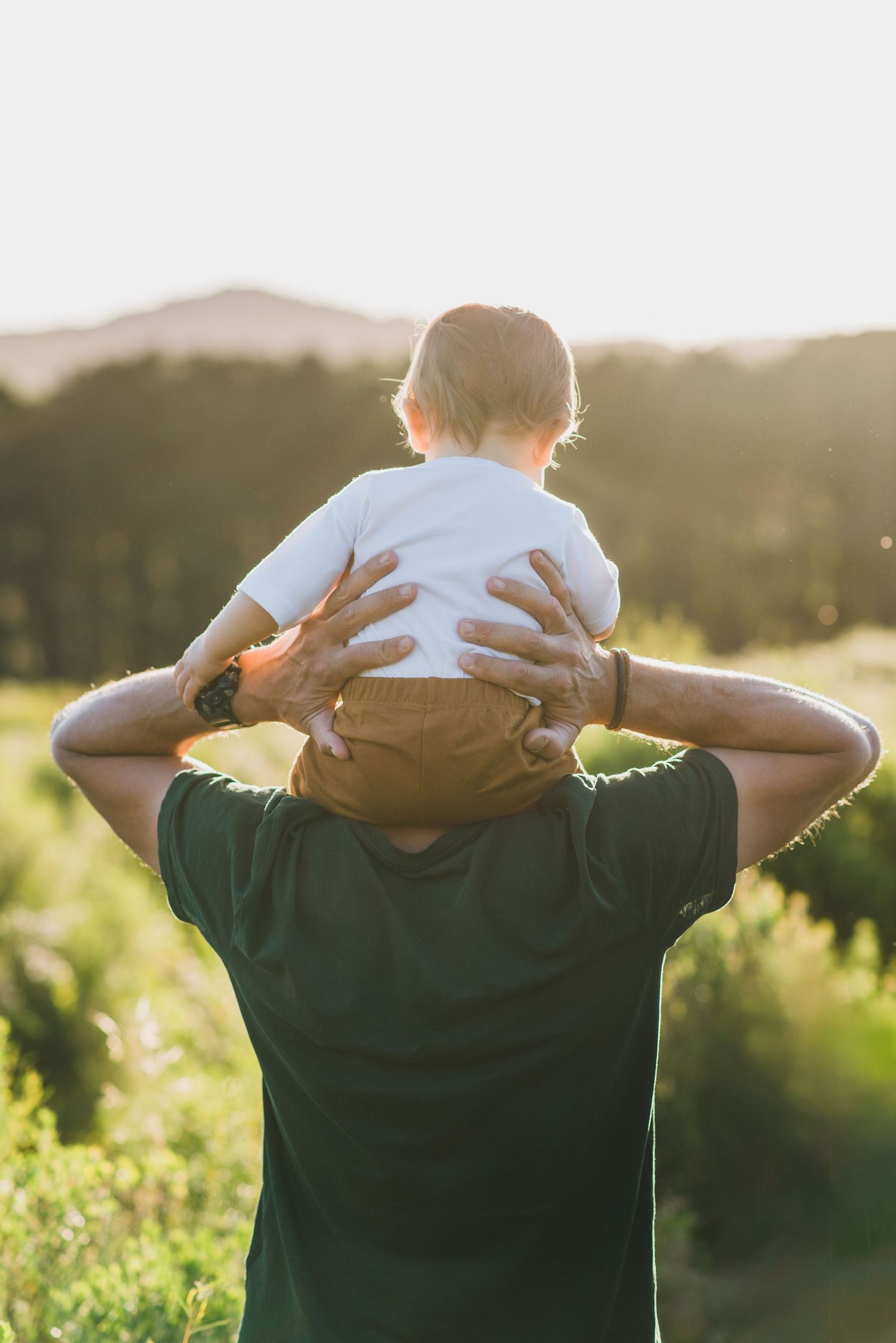 bebé de una año sobre los hombros de su padres, ambos de espalda, foto a contraluz durante el atardecer